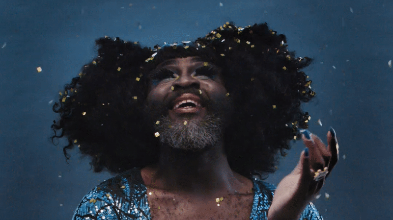 A man in drag, wearing a blue sequinned dress, watches glitter fall from the sky around him.