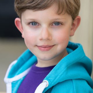 A young actor looks into the camera. They have short brown hair and are wearing a purple t shirt and a blue hoody.