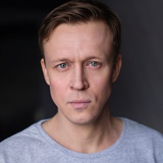 An actor looks into the camera. He has short brown hair and wears a grey t shirt.