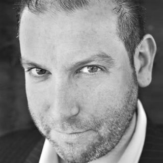 A black and white photo of an acotr looking into the camera. He wears a white shirt and dark jacket.