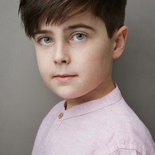 A young actor looks into the camera. He has short dark hair and wears a light pink shirt.