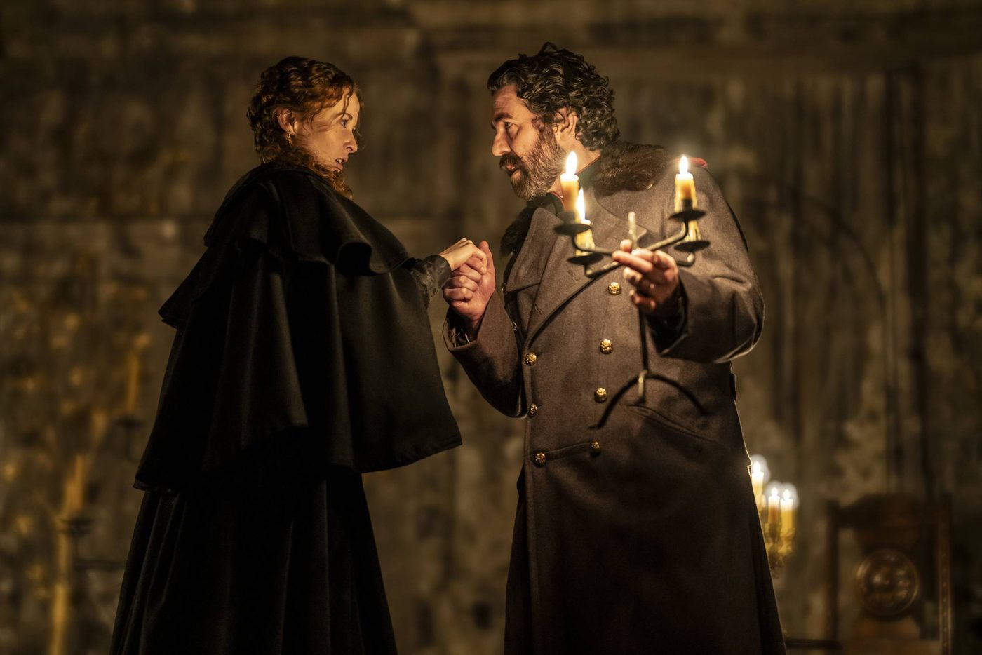 Shannon Tarbet as Masha and Paul Ready as Aleksandr Vershinin in Three Sisters. Aleksandr has grasped Masha's hand and they are looking intensely at each other. Aleksandr also holds a lit candelabra in his other hand.