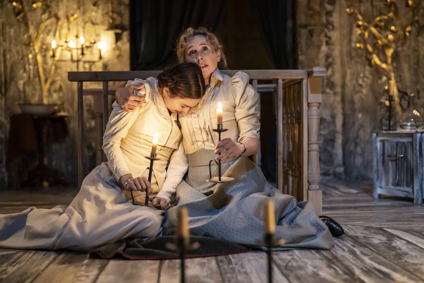 Ruby Thompson as Irina and Michelle Terry as Olga in Three Sisters. They are sat down on the floor of the stage. Irina looks as though she might be crying. Olga looks to the ceiling clutching a lit candle with one hand, the other arm draped around Irina's shoulders for comfort.