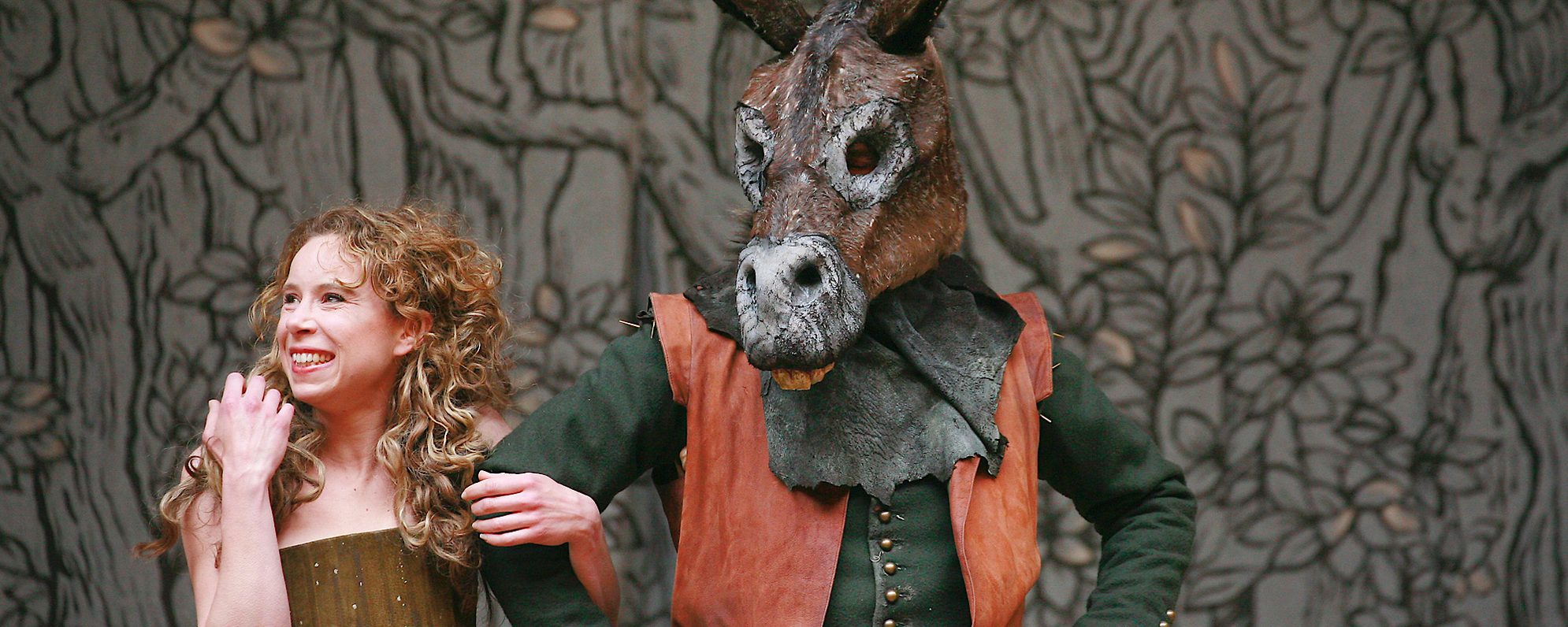 An actor smiles and holds the arm of another actor who wears an ass’ head