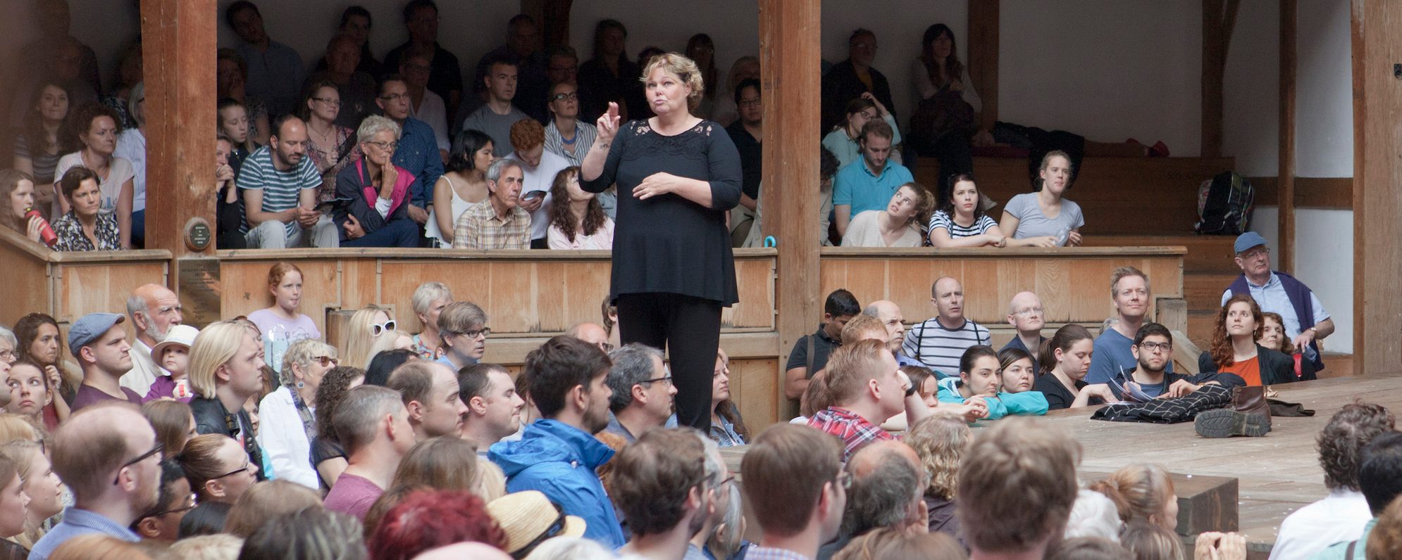A woman stands on the outdoor wooden stage of the Globe Theatre and uses her hands to sign a performance to the audience