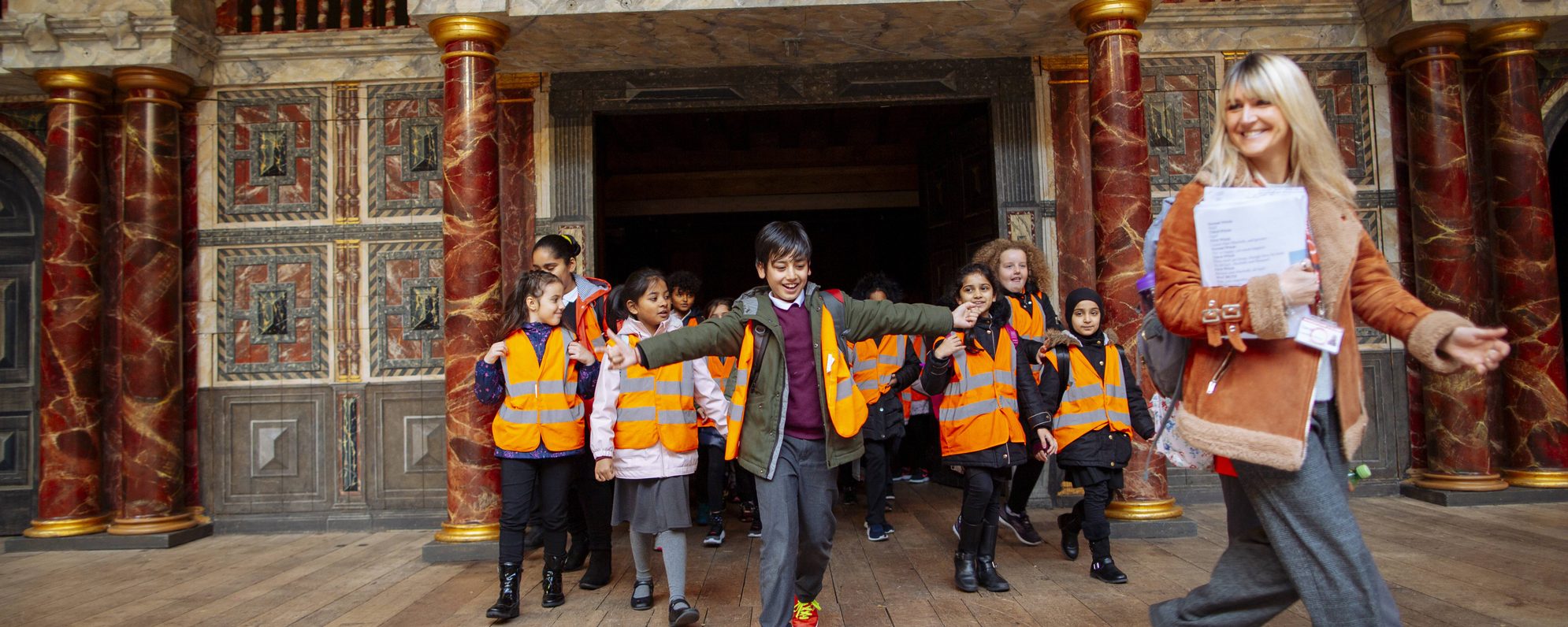 Group of primary school children in orange hi-vis vests following their teacher across the Globe stage