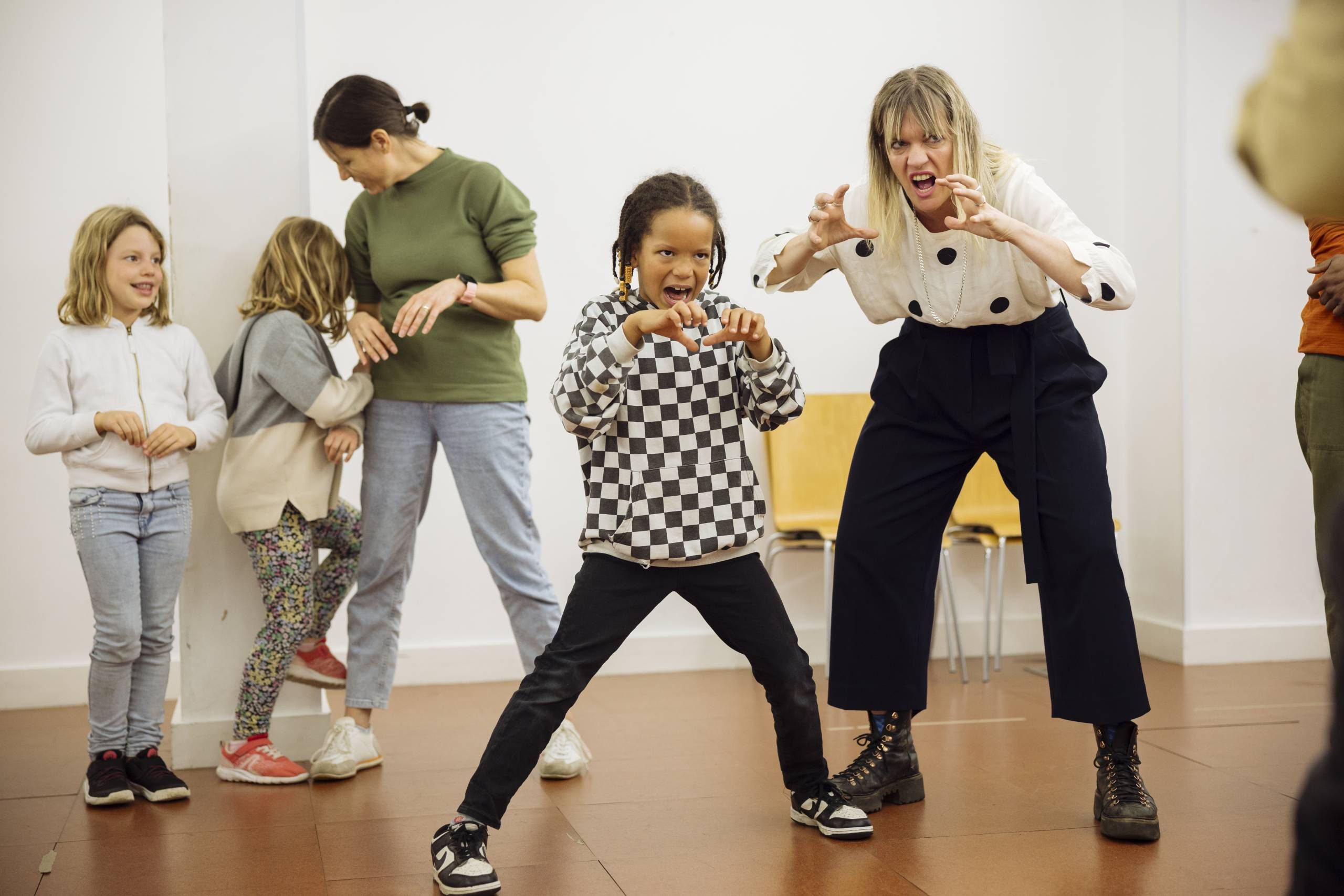 A child and parent raise their hands and expressively mime growling like an animal. In the background, two other children and a parent talk to each other. They are in a workshop room with white walls and brown flooring.