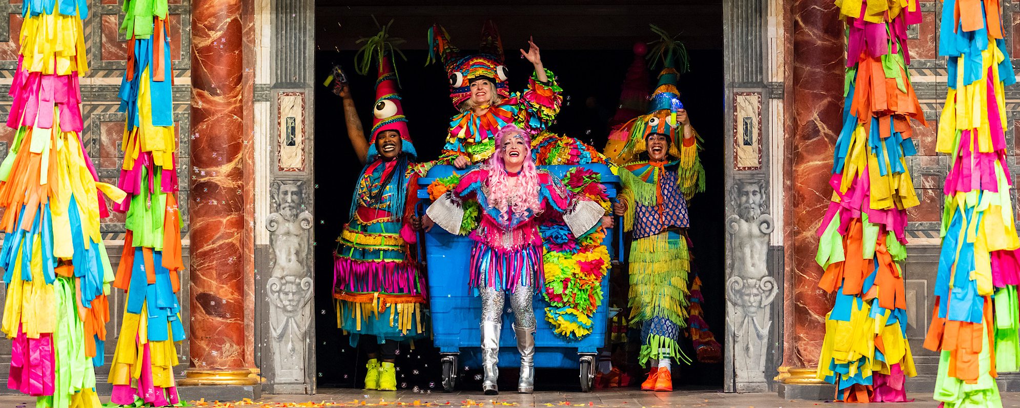 A group of actors bust onto the stage through doors, they are all dressed in bright colours and drag a blue wheelie bin which an actor sits on top of