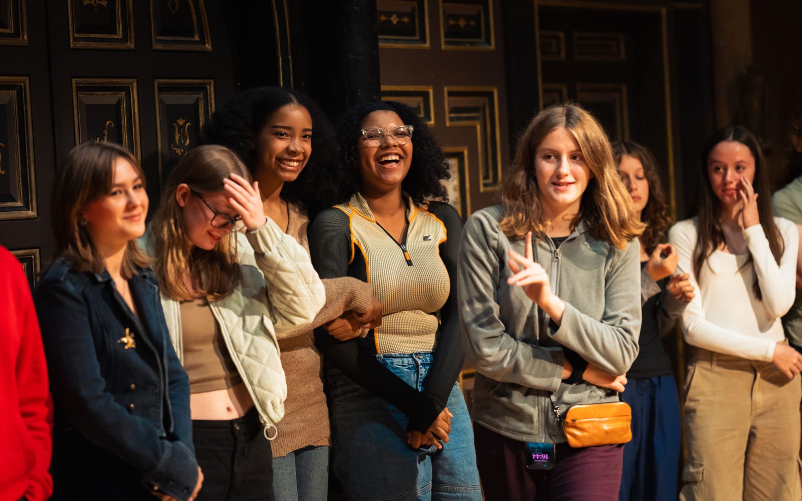 A group of young people standing in a line and smiling on the Sam Wanamaker Playhouse stage.