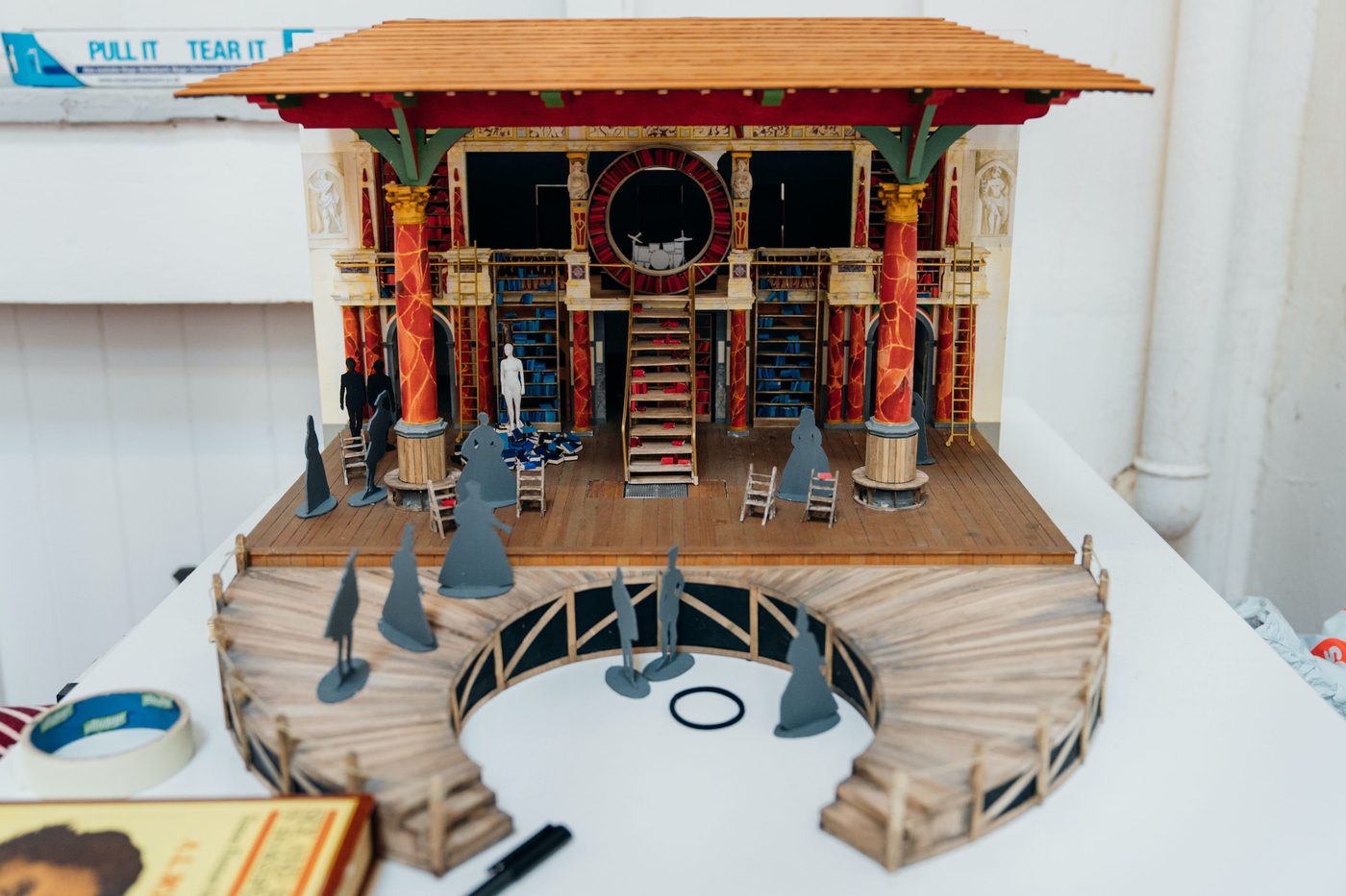 A mini set of a theatre which shows a wooden roof, wooden stage, steps, pillars and cardboard cut-out characters