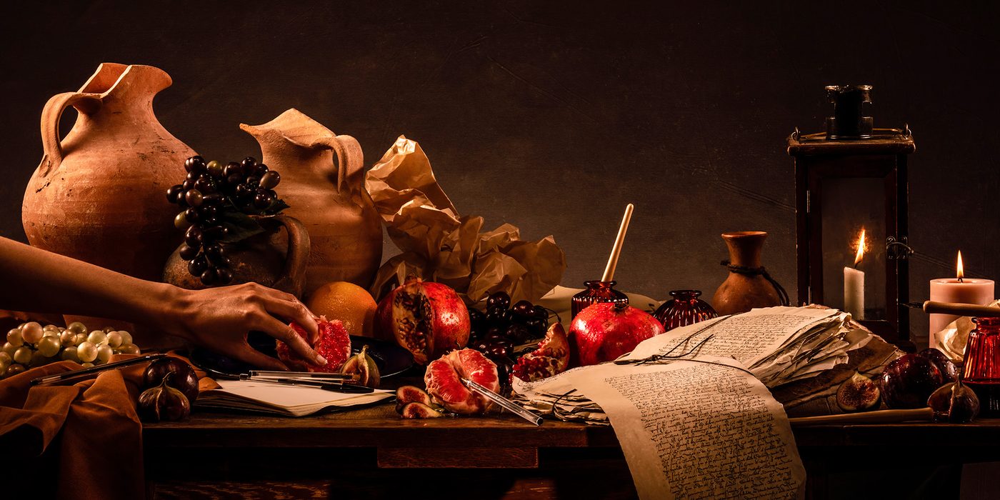 A hand reaches across a table, with jugs, food and papers spread across it.
