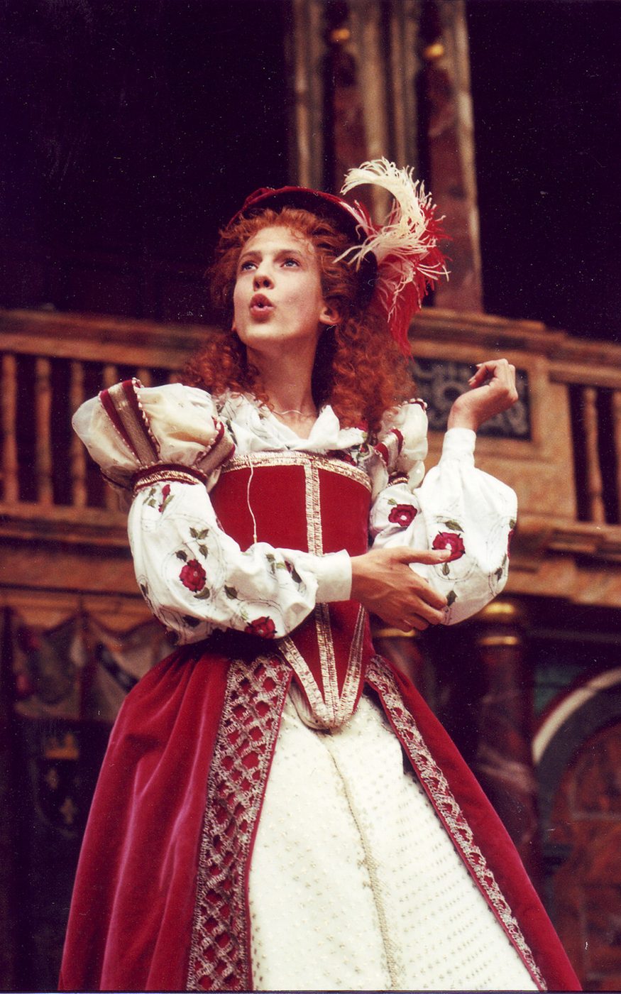 An actor wears a bright red traditional Elizabethan dress, with corset, puffed sleeves, and full skirt.