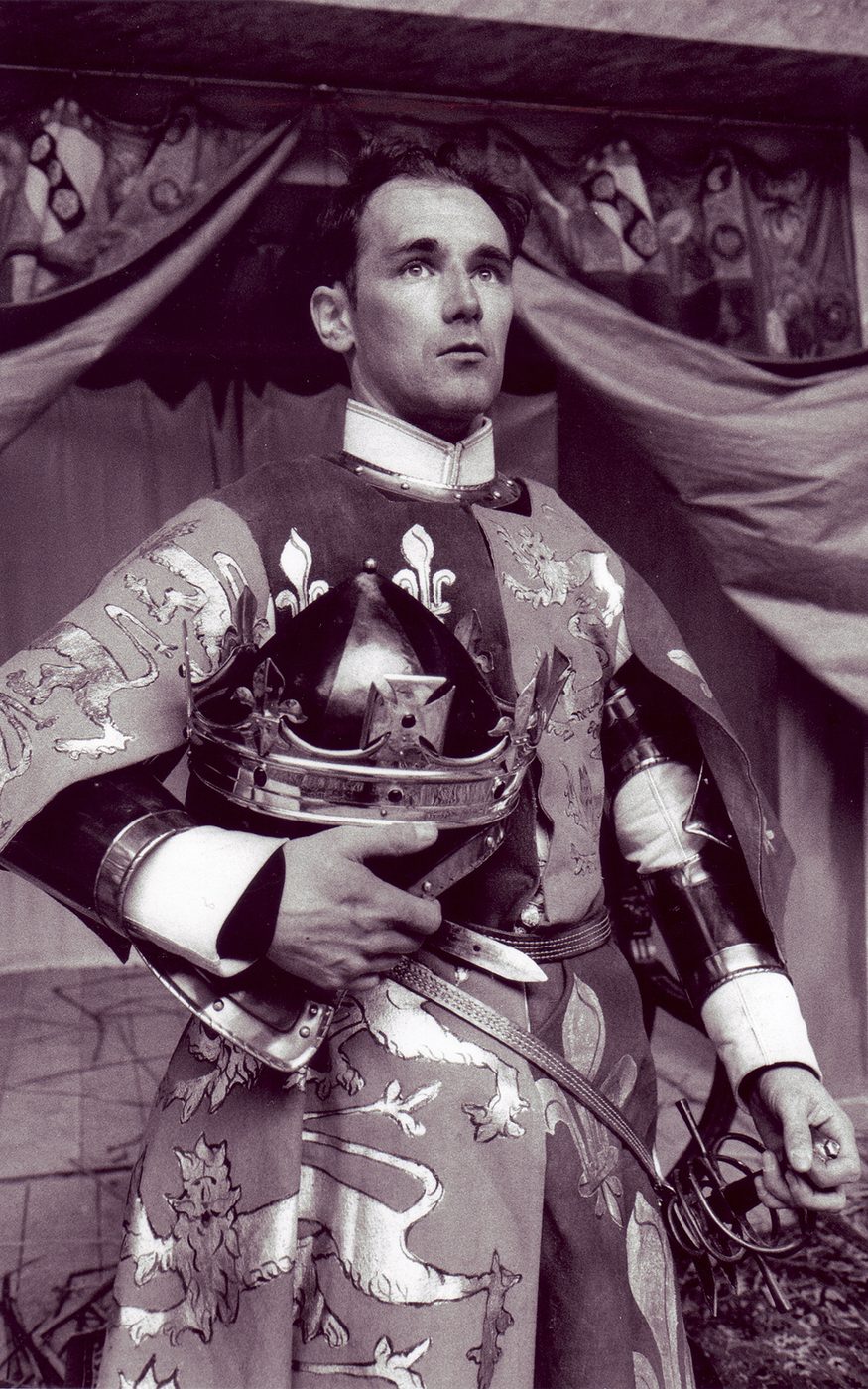 A man wearing a tunic with the English and French emblems holds his crown under his arm.