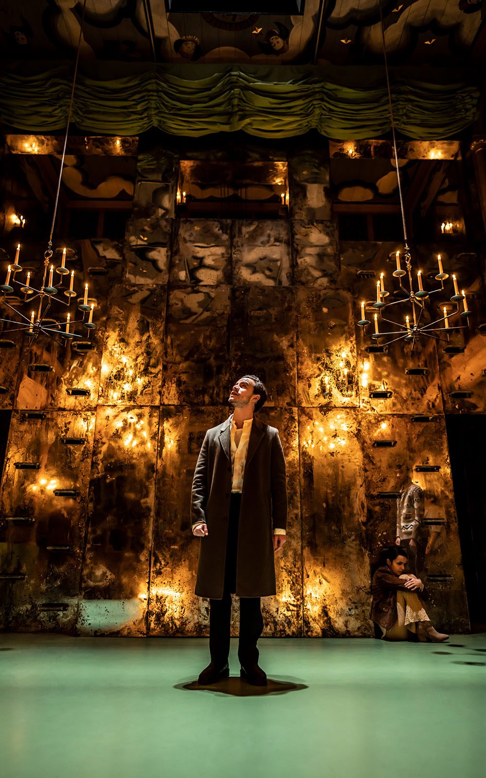 An actor stands in front of a wall of bronze on a candlelit stage