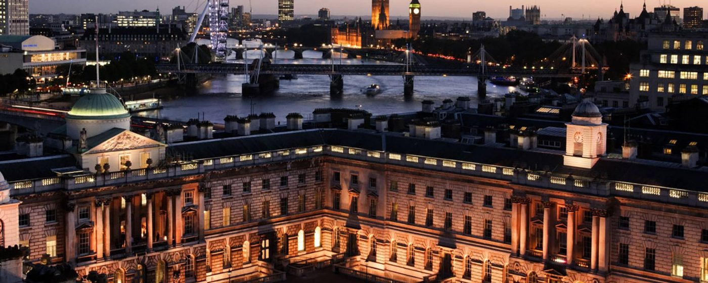 A wide nighttime shot of a large, white Neoclassical building, with the London skyline behind.