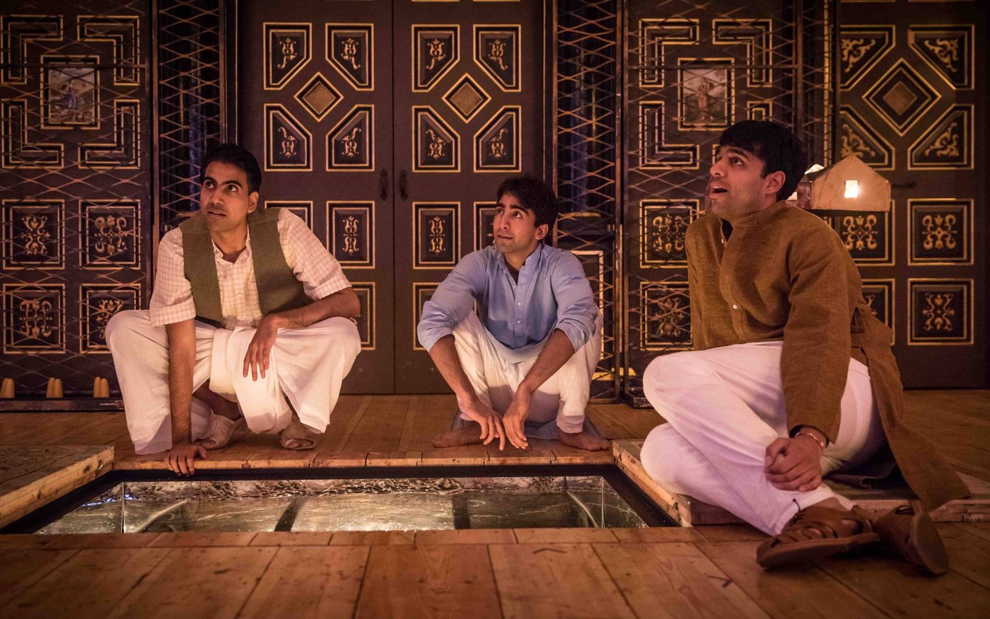Three actors sit around a trap door on a candelit stage.