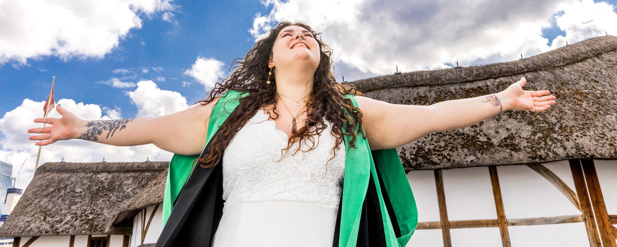 A woman wearing an academic graduation gown celebrates her MA with outstretched arms in front of the Globe.