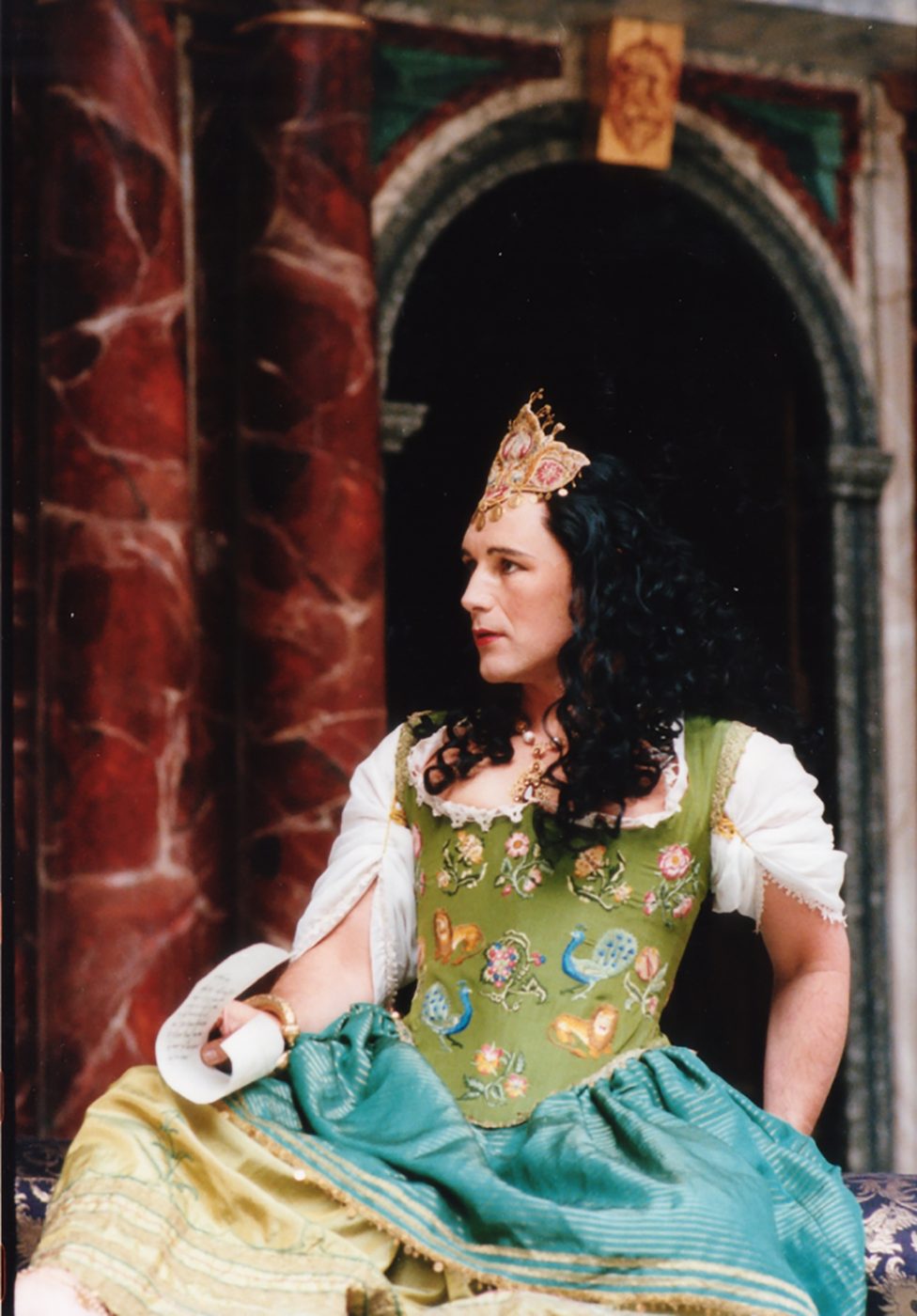 A man wearing a green corset dress sits on the Globe Theatre stage.