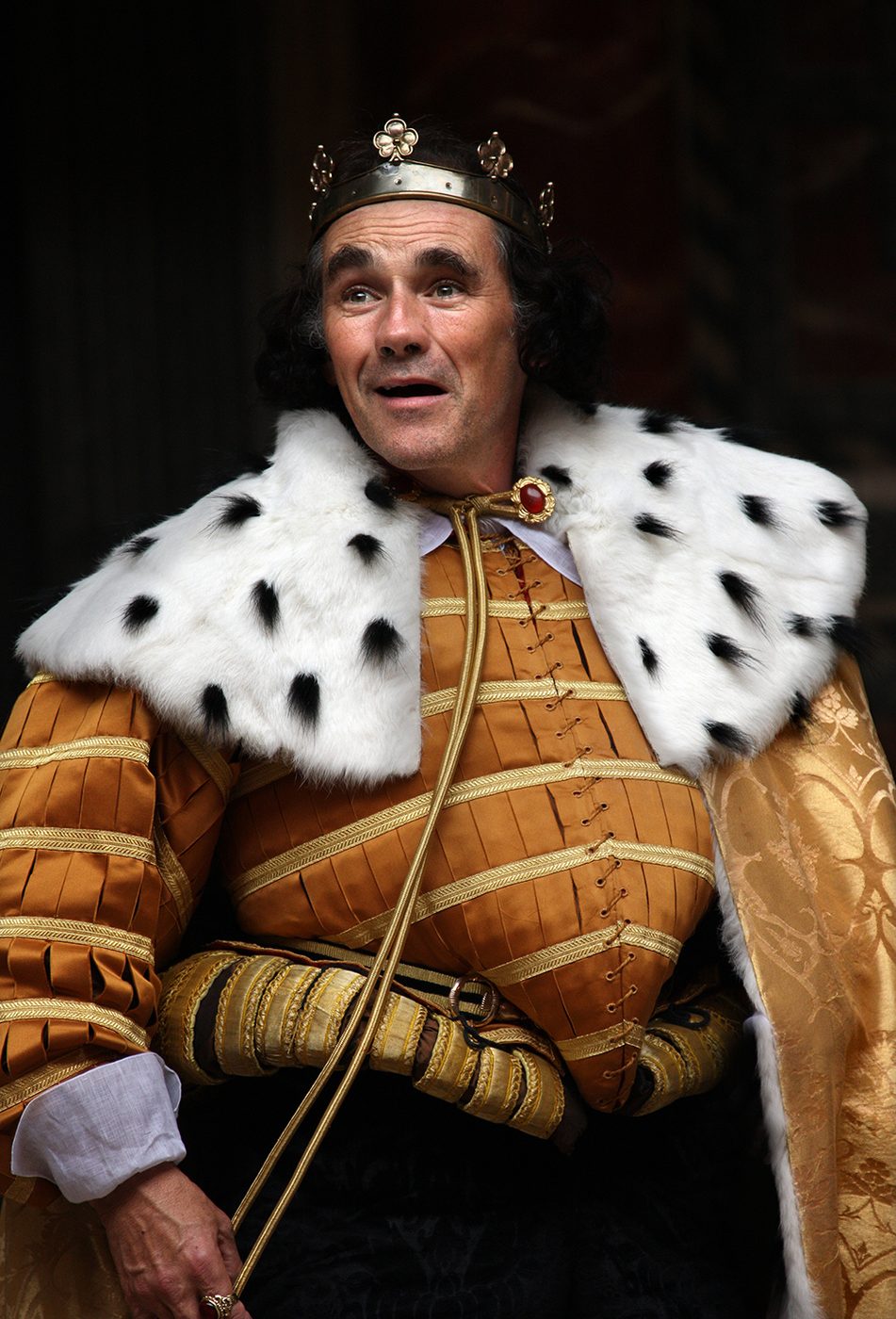 A man wearing an ermine royal cloak and golden doublet smiles.