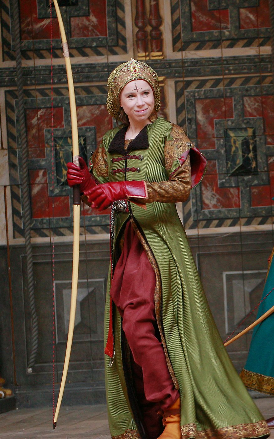 An actor wearing a green hunting coat holds a long bow.