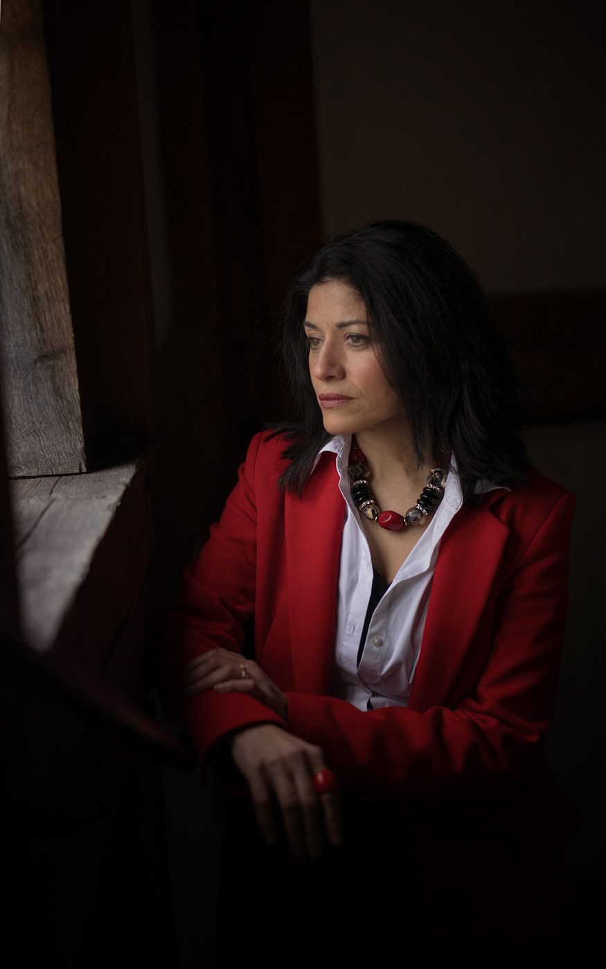 A woman wearing a red blazer looks out of a window, with daylight illuminating her.