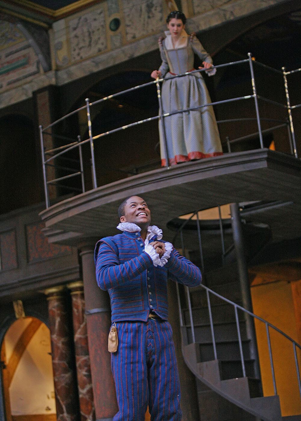 A young woman watches from a balcony as a young man clutches his hands to his chest, besotted.
