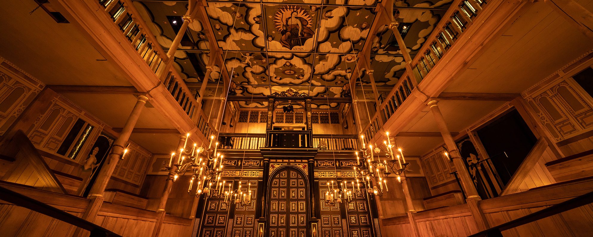 A wide angled shot of the Sam Wanamaker Playhouse with its ornate painting ceiling depicting the Goddess Luna and clouds, hanging candelabra, and panelled painted doors.