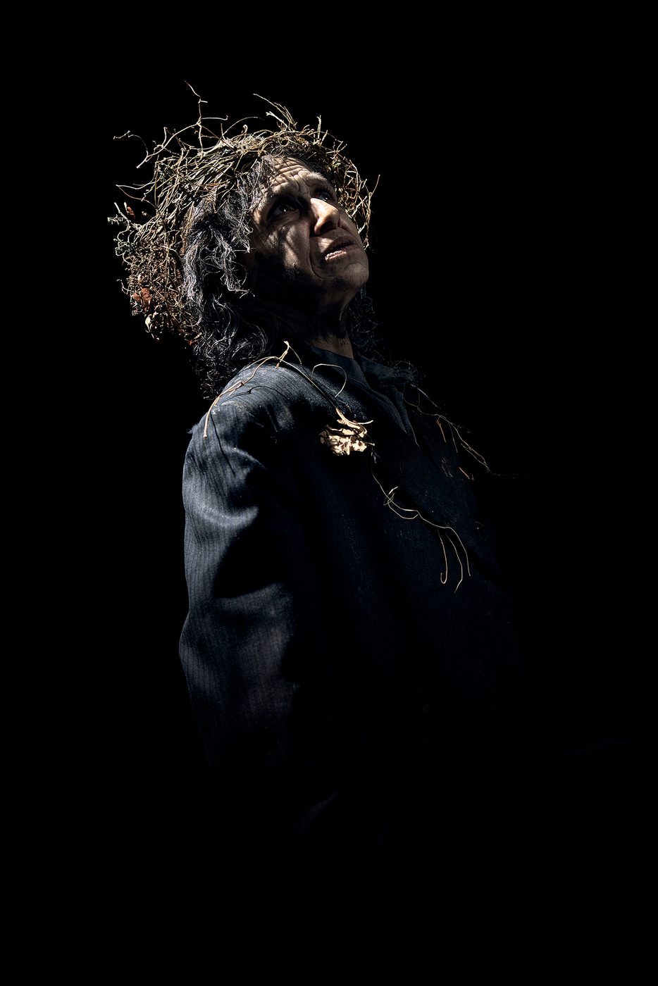 An actor wearing a wreath crown of branches and leaves looks up, their face cast in shadow.