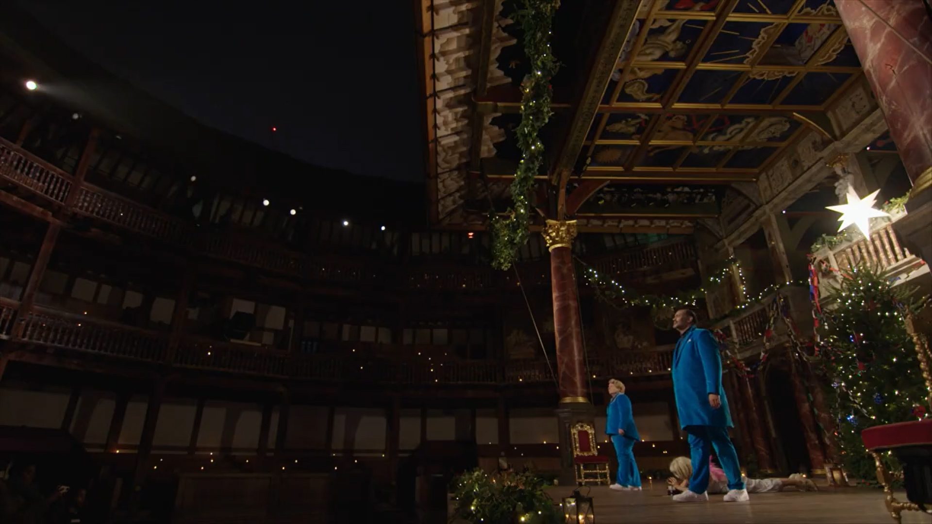 Two actors wearing matching blue suits step out onto the Globe Theatre stage.