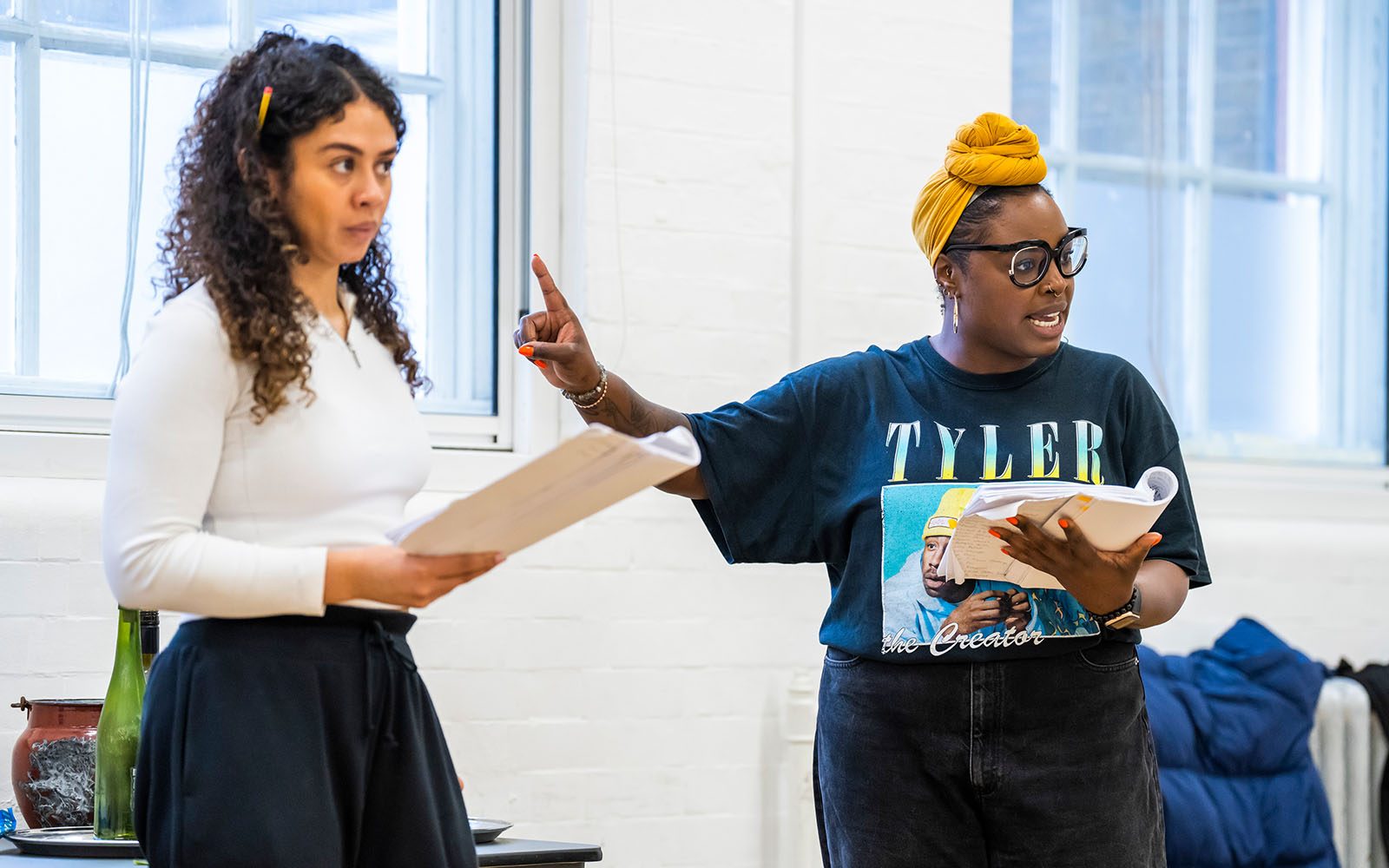 Two actors are standing in a rehearsal room holding cue scripts. One of them holds her finger up to the other, who is looking sideways at her.