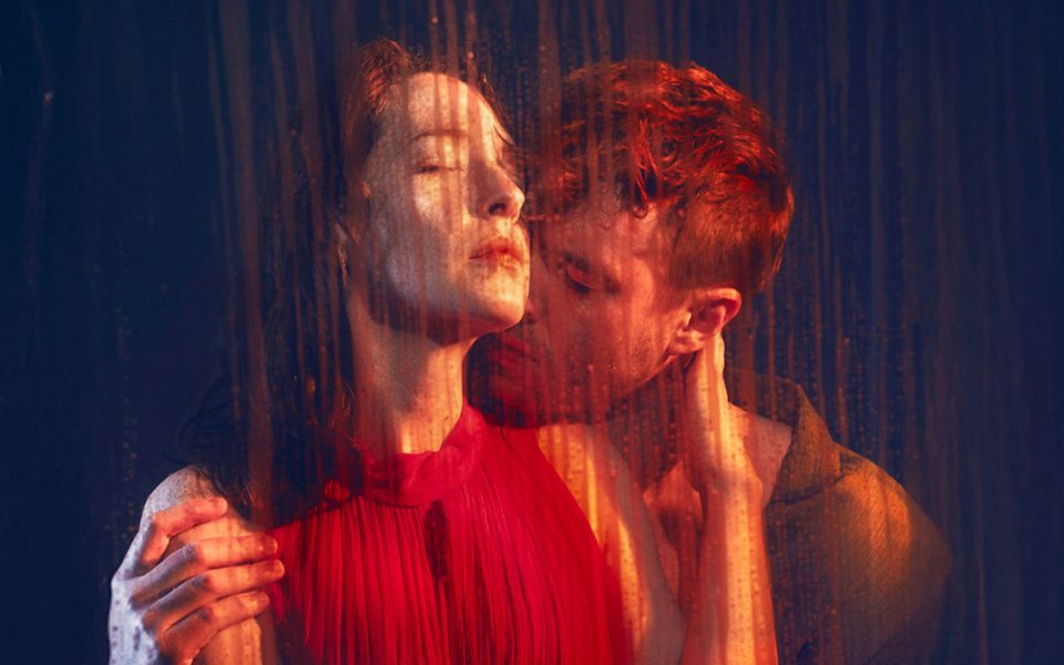 Two people embrace in front of a dark blue backdrop, with droplets of water running down a pane of glass in front of them.