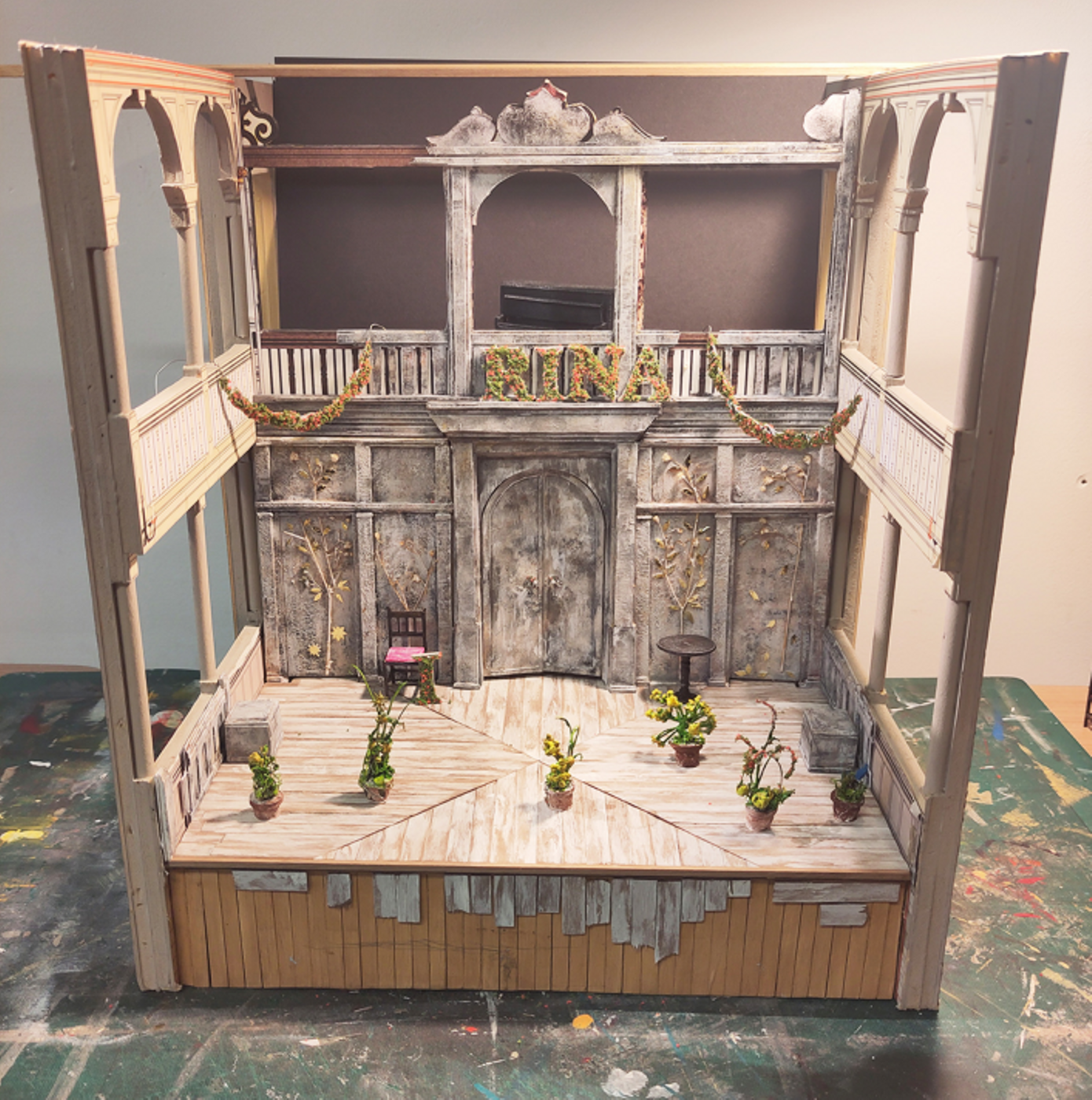 Model box set design of Three Sisters by Oli Townsend. The model shows a relatively bare set. The walls and floor of the stage have been clad in silver birch, and on the back wall there are four silver birch trees depicted, with gold leaves. A floral arrangement spells out 