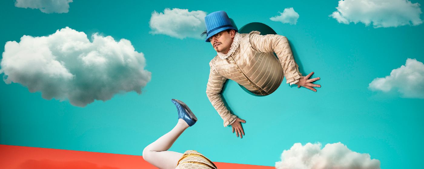 The torso of a man in Elizabethan dress appears from a hole in a blue backdrop with clouds. His legs appear from a different hole in the ground