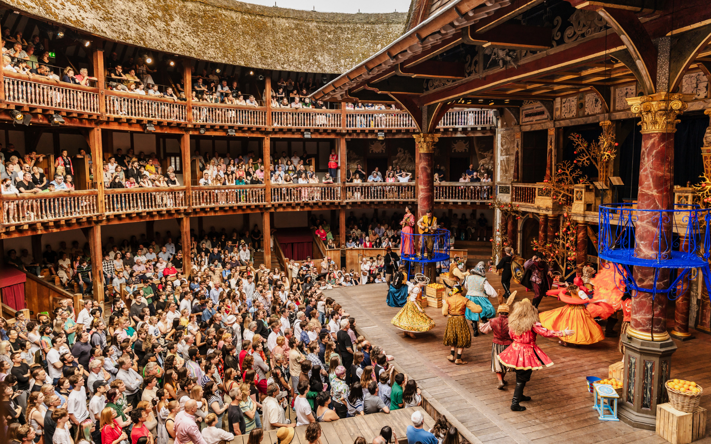 A photograph of the Globe Theatre during a performance. On stage are a group of actors dancing in colourful costumes. The auditorium is full of people standing and sitting.